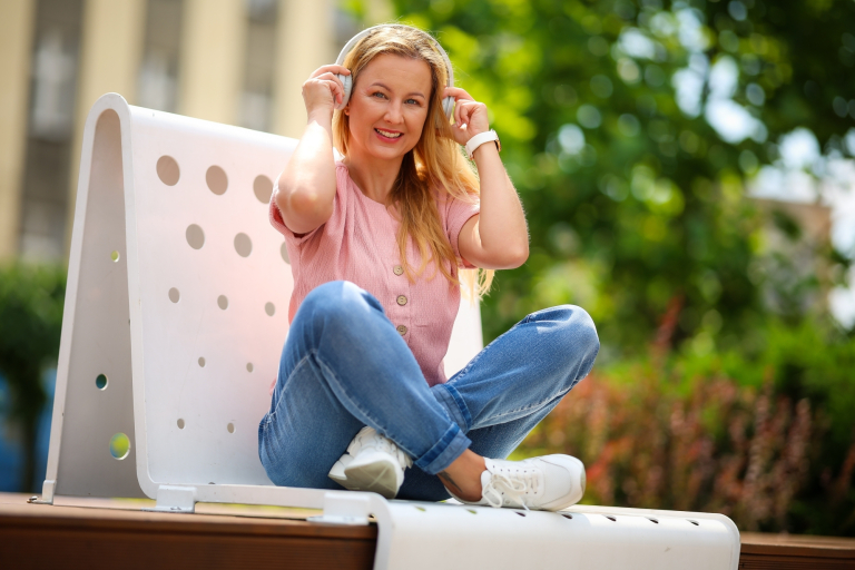 Strona główna | Uniwersytet WSB Merito Chorzów: Studentka WSB Merito siedzi na ławce w plenerze i słucha muzyki przez słuchawki