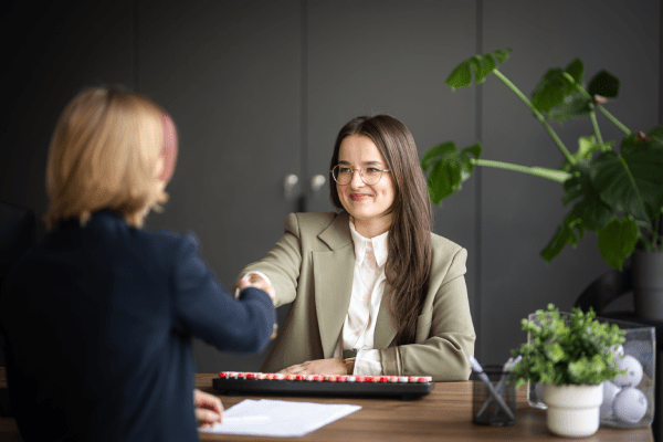IACBE Accreditation | WSB Merito Gdańsk University: Dwie kobiety podają sobie rękę w geście zawarcia umowy. Jedna z nich siedzi za biurkiem, przy klawiaturze, uśmiecha się. Ma na sobie okulary i marynarkę o delikatnym odcieniu zieleni. Tło jest ciemne, ale wyraźnym elementem jest duża zielona monstera. 