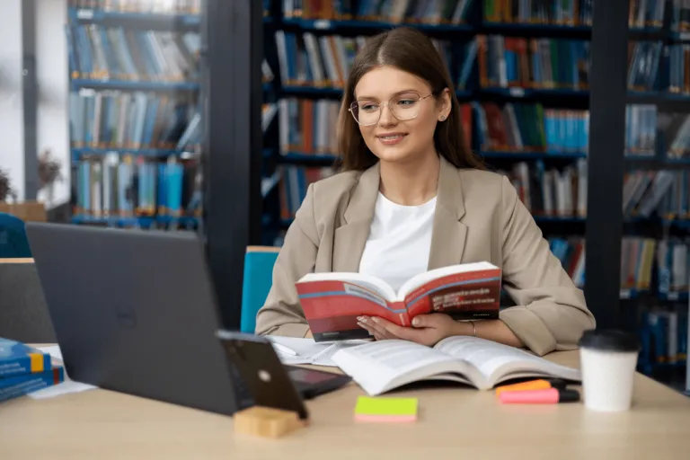 Digital university | WSB Merito Gdańsk University: Zdjęcie studentki w okularach siedzącej przed laptopem z książką. Znajduje się ona najprawdopodobniej w bibliotece, ponieważ z tyłu są zapełnione regały księgozbiorem. Jest ona w stroju biznesowym