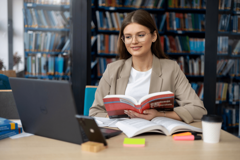 Digital university | WSB Merito Gdańsk University: Zdjęcie studentki w okularach siedzącej przed laptopem z książką. Znajduje się ona najprawdopodobniej w bibliotece, ponieważ z tyłu są zapełnione regały księgozbiorem. Jest ona w stroju biznesowym