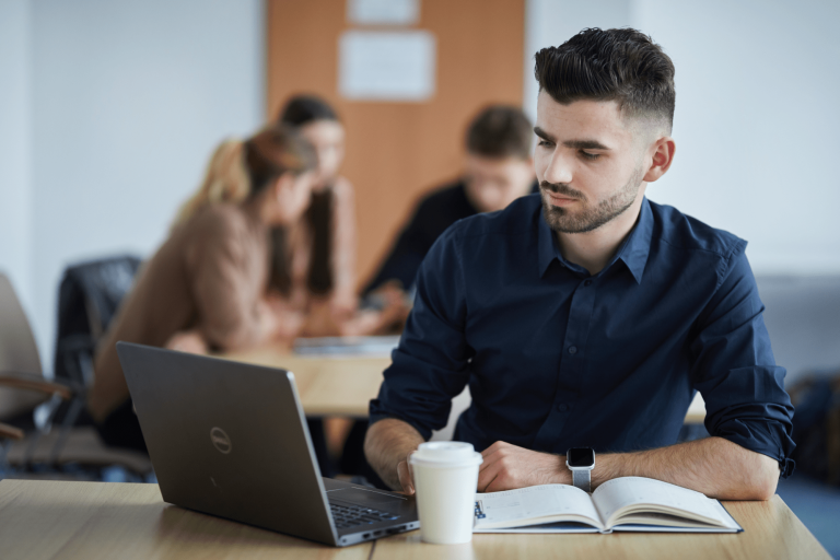 Digital university | WSB Merito Gdańsk University: Młody mężczyzna w ciemnej koszuli siedzi przy laptopie. Siedzi w klasie, w tle widać grupę rozmawiających studentek