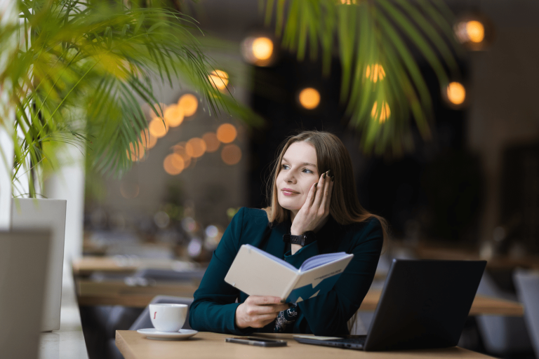 Library | WSB Merito Gdańsk University: Portret młodej, naturalnej kobiety o delikatnych rysach i włosach. Siedzi przy biurku w kawiarni oparta dłonią o głowę, w drugiej ręce trzyma zeszyt z logotypem UWSB Merito