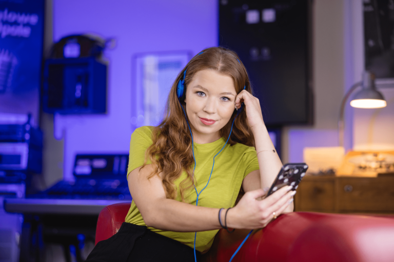 Digital university | WSB Merito Gdańsk University: Na zdjęciu znajduje się młoda dziewczyna, która ma w ręku komórkę oraz nosi niebieskie słuchawki. Ma ona rudawe włosy i lekko się uśmiecha