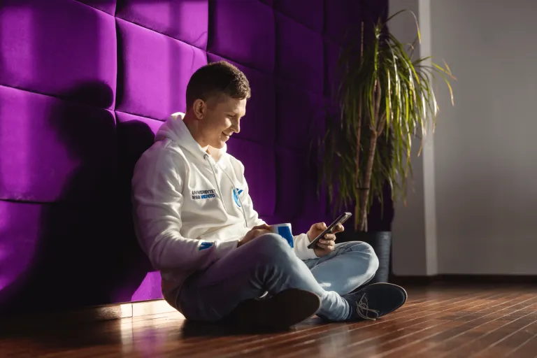 Digital university | WSB Merito Gdańsk University: Zdjęcie studenta siedzącego na podłodze i opierającego się na fioletowej miękkiej ścianie. Przegląda on komórkę i ma w ręku kubek. Uśmiecha się, nosi białą bluzę z logo Uniwersytetu WSB Merito