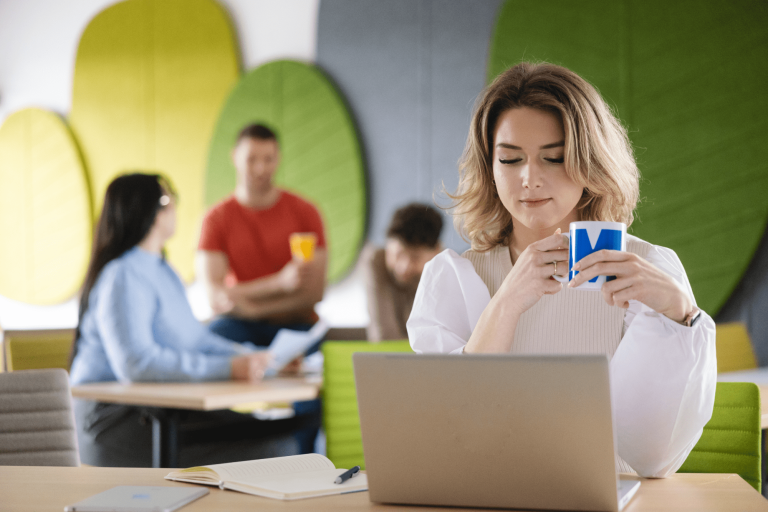 Digital university | WSB Merito Gdańsk University: Zdjęcie przedstawia osoby znajdujące się w przestrzeni, gdzie na ścianie znajdują się materiałowe liście. Siedzą przy stolikach. Na głównym planie jest kobieta przy laptopie, trzyma w ręku kubek