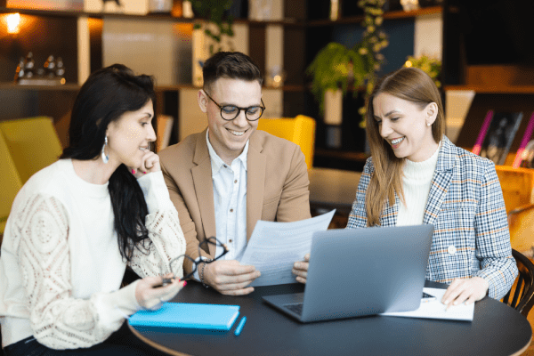 Research Federation | WSB Merito Gdańsk University: Dwie kobiety oraz mężczyzna w eleganckich strojach dyskutują nad wspólną pracą w przestrzeni z ciepłym światłem. Siedzą przed stolikiem i laptopem