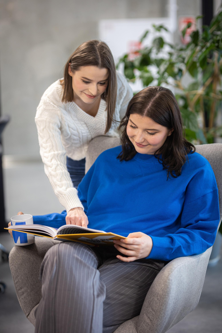 Library | WSB Merito Gdańsk University: Studentka siedząca na fotelu z książką i pochylająca się nad nią dziewczyna i pokazująca coś w publikacji