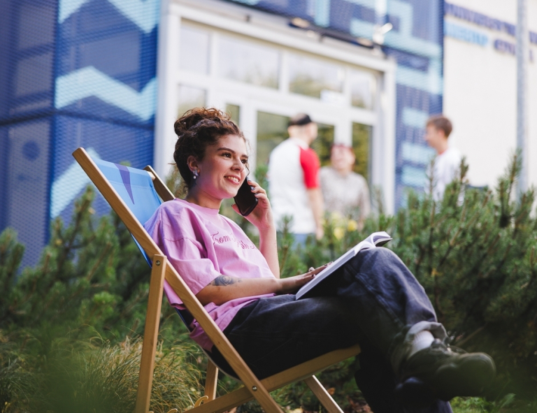Digital Marketing, studia II stopnia, magisterskie: Studentka siedzi na leżaku przed kampusem z komórką w ręku i książką leżącą na kolanach