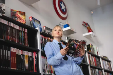 Student organisations and research clubs | WSB Merito Łódź University: Kobieta w niebieskiej koszuli czyta komiks, otoczona półkami pełnymi komiksów i dekoracjami Marvela.