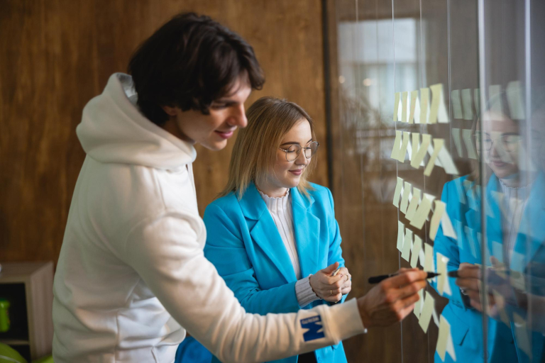 Management, studia I stopnia, licencjackie i inżynierskie: Dwoje studentów pracuje wspólnie przy szklanej tablicy z karteczkami, planując i omawiając elementy projektu.