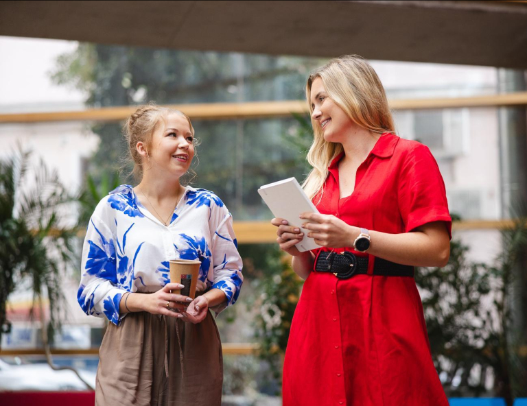 University without barriers | WSB Merito Opole University: Dwie rozmawiające dziewczyny. Blondynka w jasnej koszuli trzyma kubek kawy, druga dziewczyna w czerwonej sukience trzyma notatnik 