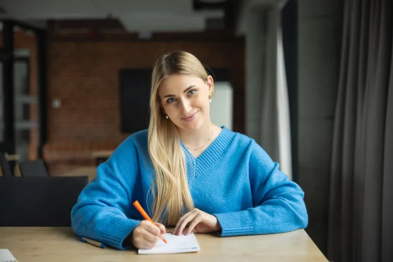 Strategic Management | WSB Merito Poznań University: kobieta w niebieskim swetrze siedzi przy stole i z uśmiechem sporządza notatki