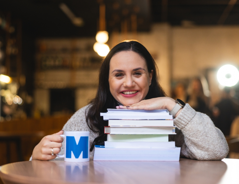 Search results | WSB Merito Universities: Uśmiechnięta kobieta przy stole opierająca się na stosie książek z kubkiem Merito w dłoni