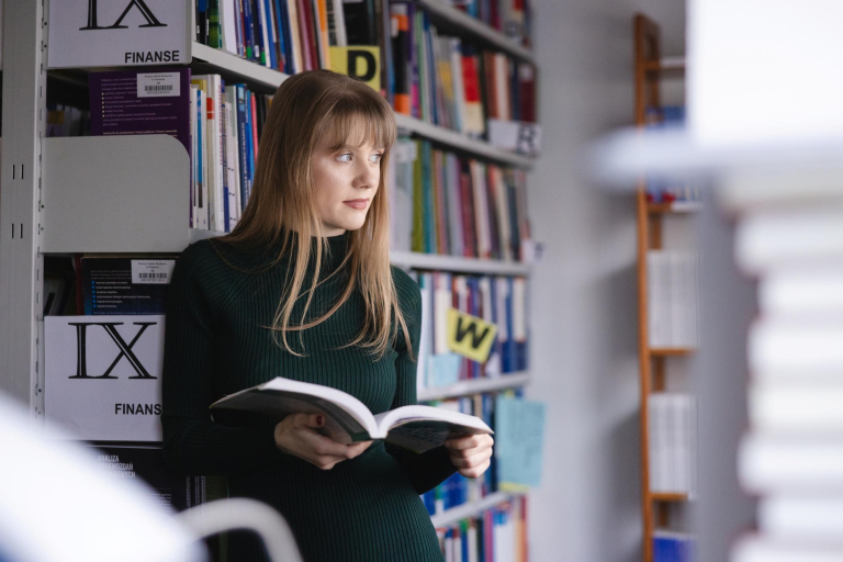 Social impact | Uniwersytet WSB Merito Szczecin: Studentka Uniwersytetu WSB Merito spogląda za okno, przeglądając książkę w bibliotece.