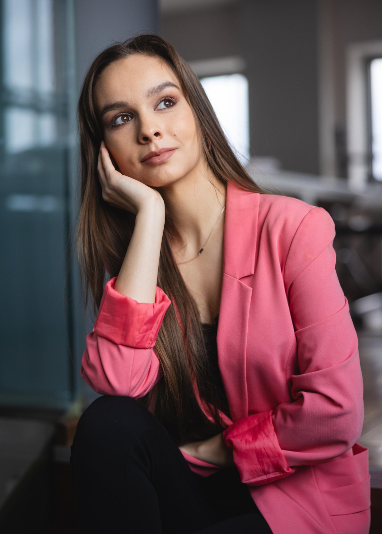 Who we are | WSB Merito Toruń University: studentka z długimi włosami i w marynarce w kolorze łososiowym, siedzi na sofie, patrzy za okno, głowę podpiera na ręce