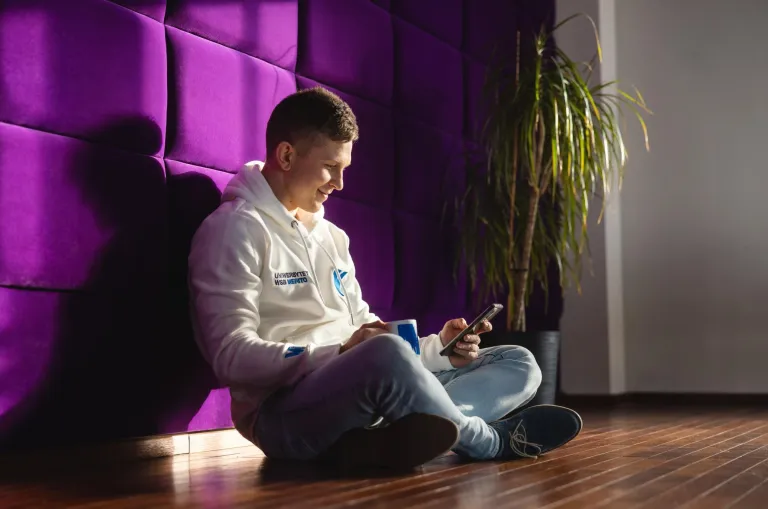 student siedzi na podłodze ze skrzyżowanymi nogami, telefonem i kubkiem w ręku