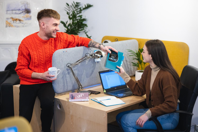 Who we are | WSB Merito Wrocław University: Kobieta i mężczyzna siedzą przy biurkach i podają sobie zeszyt