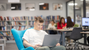 Bezpieczeństwo wewnętrzne | Studia II stopnia ze studiami podyplomowymi w Gdańsku | Uniwersytet WSB Merito w Gdańsku: Student siedzi na fotelu z laptopem na kolanach w bibliotece