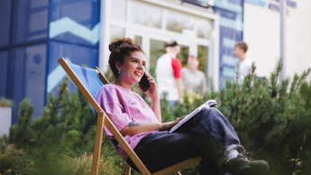 Administrator baz danych | Studia I stopnia w Gdańsku | Uniwersytet WSB Merito w Gdańsku: Studentka siedzi na leżaku przed kampusem z komórką w ręku i książką leżącą na kolanach
