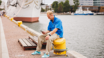 Biały wywiad | Studia I stopnia w Gdańsku | Uniwersytet WSB Merito w Gdańsku: Student siedzi na kołku i przegląda komórkę w tle widać statek