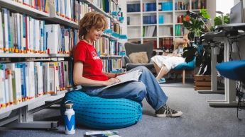 Terapia pedagogiczna z elementami integracji sensorycznej | Studia II stopnia ze studiami podyplomowymi  | Uniwersytet WSB Merito w Gdańsku: Studentka w bibliotece siedzi na puffie i przegląda książkę 