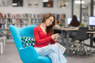 Biblioteka | Uniwersytet WSB Merito w Gdańsku: Studentka w bibliotece siedzi na wygodnym fotelu i korzysta z komórki