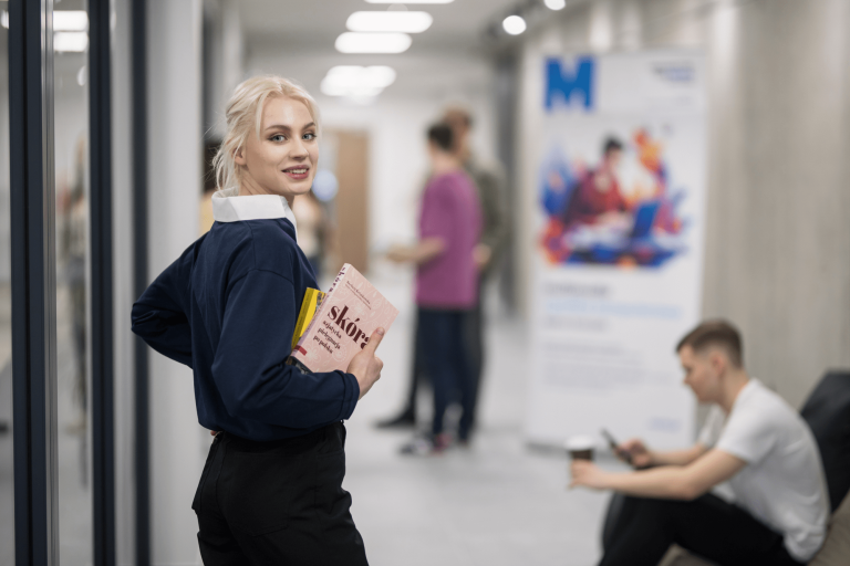 ABC STUDENTA - szkolenie - do zmodyfikowania | Uniwersytet WSB Merito w Gdańsku: Zdjęcie studentki o jasnych blond włosach - patrzy się ona w obiektyw i trzyma książkę. W tle studenci na korytarzu