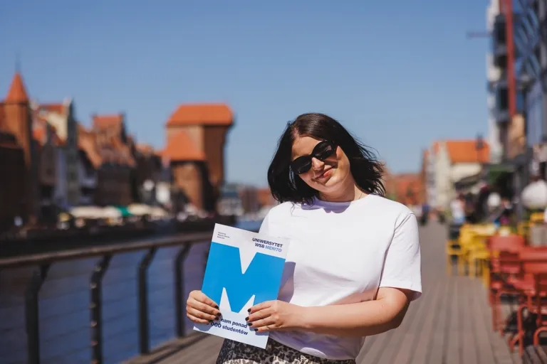 Czesne, stypendia i zniżki | Uniwersytet WSB Merito w Gdańsku: Studentka z teczką z sygnetem M, z tyłu widać Gdańskiego Żurawia