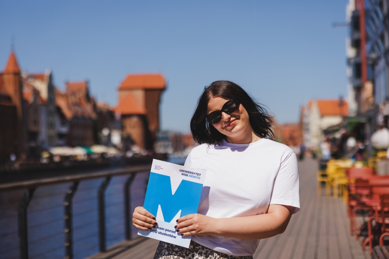 Czesne, stypendia i zniżki | Uniwersytet WSB Merito w Gdańsku: Studentka z teczką z sygnetem M, z tyłu widać Gdańskiego Żurawia