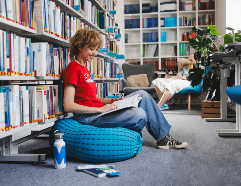 Zarządzanie projektami i usługami IT | Studia II stopnia ze studiami podyplomowymi  | Uniwersytet WSB Merito w Gdańsku: Studentka w bibliotece siedzi na puffie i przegląda książkę 