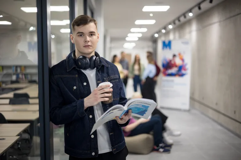 Zarządzanie zasobami ludzkimi | Studia II stopnia w Gdańsku | Uniwersytet WSB Merito w Gdańsku: Student z książką i kawą stoi opierając się o szklaną ścianę na korytarzu na Uniwersytecie 