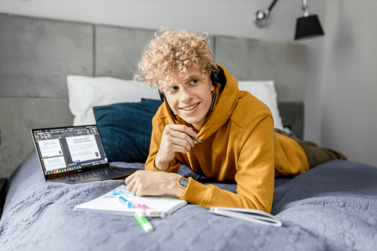 ESG w systemach bezpieczeństwa i higieny pracy | Studia I stopnia w Gdańsku | Uniwersytet WSB Merito w Gdańsku: Student leży na łóżku z notatkami i laptopem