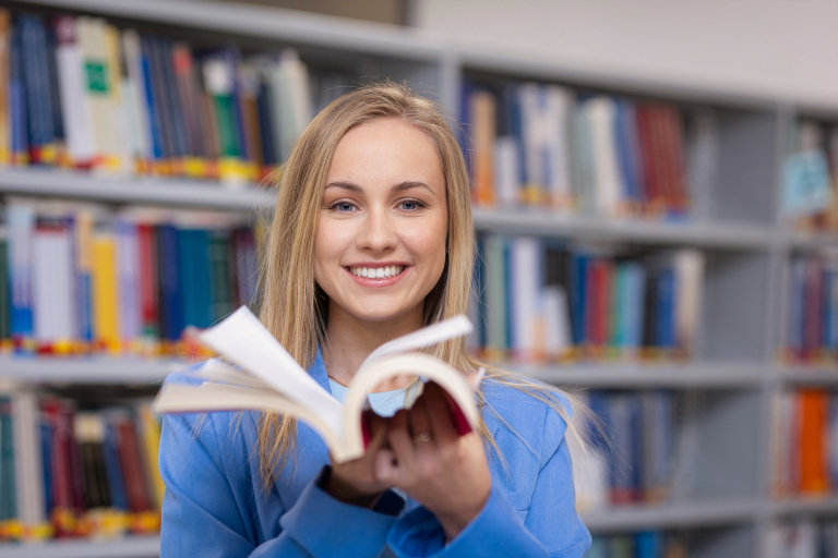 Strefa studenta | Uniwersytet WSB Merito w Gdańsku: Uśmiechnięta studentka WSB Merito wertuje książkę w bibliotece