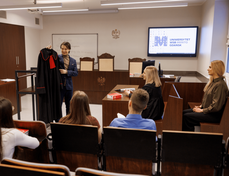 Prawo | Studia jednolite magisterskie w Gdańsku | Uniwersytet WSB Merito w Gdańsku: Warsztaty na sali sądowej, jedna z osób pokazuje togę