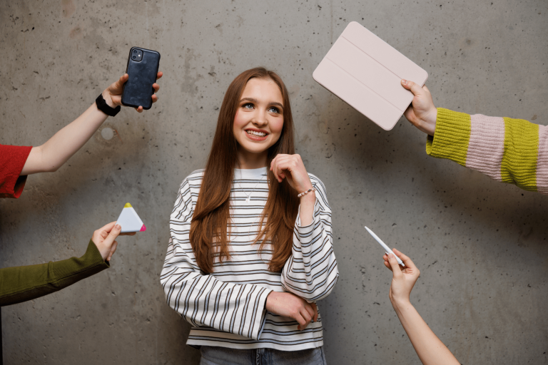 Program Very Important Student | Uniwersytet WSB Merito w Gdańsku: Studentka na kreatywnym portrecie - stoi przy ścianie, a wokół niej dłonie z przedmiotami: tablet, komórka, mazak, długopis
