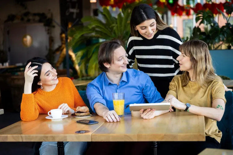 Tłumaczenia - język angielski | Uniwersytet WSB Merito w Gdańsku: Studenci siedzą przy biurku w kawiarni i rozmawiają, jedna z osób trzyma tablet