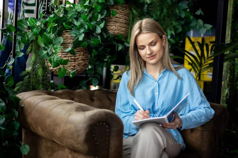 Terapia pedagogiczna z elementami integracji sensorycznej | Studia II stopnia ze studiami podyplomowymi  | Uniwersytet WSB Merito w Gdańsku: Studentka siedzi w fotelu i robi notatki. Ma na sobie jasną niebieską koszulę. Wokół niej jest zielona roślinność