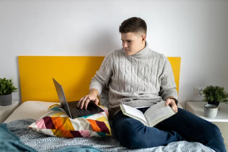 Zarządzanie projektami i usługami IT | Studia I stopnia w Gdańsku | Uniwersytet WSB Merito w Gdańsku: Student siedzi w łóżku z laptopem i ma na kolanach książkę