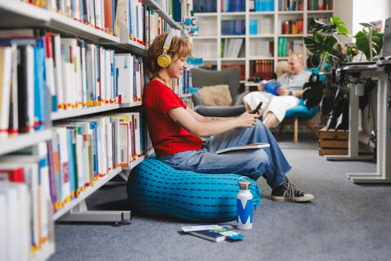 Wyniki wyszukiwania | Uniwersytet WSB Merito w Gdańsku: Studentka z książką na kolanach, słuchawkami na uszach i z komórką w ręku we wnętrzu biblioteki