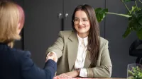 Zarządzanie nieruchomościami i pośrednictwo | Studia I stopnia w Gdyni | Uniwersytet WSB Merito Gdynia: Zdjęcie przedstawia sytuację biznesową. Obie kobiety siedzące przy biurkach podają sobie ręce - w domyśle po podpisaniu umowy. Jedną kobietę widać z frontu - uśmiecha się i ma widoczny dołeczek. Ma na sobie oliwkową marynarkę. Z tyłu znajduje się ciemna szara ściana, którą ociepla trochę duża monstera 