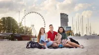 Kontynuuj studia | Uniwersytet WSB Merito Gdynia: Fotografia robiona na plaży w Gdyni, z tyłu widać wieżowiec i koło widokowe. Na plaży siedzą dwie studentki i student