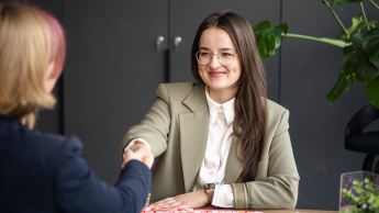 Zarządzanie nieruchomościami i pośrednictwo | Studia I stopnia w Gdyni | Uniwersytet WSB Merito Gdynia: Zdjęcie przedstawia sytuację biznesową. Obie kobiety siedzące przy biurkach podają sobie ręce - w domyśle po podpisaniu umowy. Jedną kobietę widać z frontu - uśmiecha się i ma widoczny dołeczek. Ma na sobie oliwkową marynarkę. Z tyłu znajduje się ciemna szara ściana, którą ociepla trochę duża monstera 