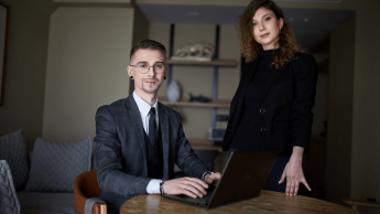 Zdjęcie osób w średnim wieku. Mężczyzna w garniturze siedzi przy laptopie, obok niego stoi kobieta ubrana na czarno