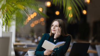 Język angielski w biznesie | Studia I stopnia w Gdyni | Uniwersytet WSB Merito Gdynia: Portret młodej, naturalnej kobiety o delikatnych rysach i włosach. Siedzi przy biurku w kawiarni oparta dłonią o głowę, w drugiej ręce trzyma zeszyt z logotypem UWSB Merito 