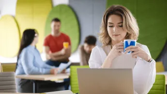 Pedagogika resocjalizacyjna z profilaktyką społeczną | Studia I stopnia w Gdyni | Uniwersytet WSB Merito Gdynia: Zdjęcie kobiety siedzącej w przestrzeni kawiarni przy laptopie. Z tyłu widać innych ludzi. Ma ona na sobie jasne ubrania i trzyma kubek z logo Merito