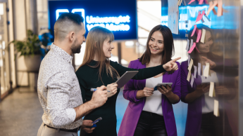 Zdjęcie przedstawiające dwie studentki i studenta pracującego w grupie. Stoją oni przy karteczkach i coś omawiają. Ubrani są biznesowo. Z tyłu widać neon Uniwersytetu WSB Merito