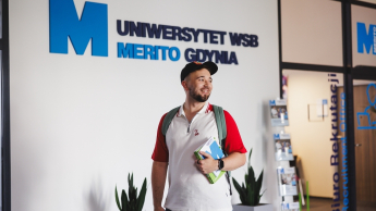 Kadry i płace | Studia I stopnia w Gdyni | Uniwersytet WSB Merito Gdynia: Student na korytarzu przy logo Uczelni
