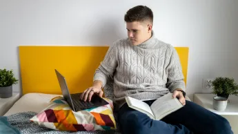 Finanse i rachunkowość | Studia I stopnia w Gdyni | Uniwersytet WSB Merito Gdynia: Uczeń z książką na kolanach siedzi w łóżku przy laptopie