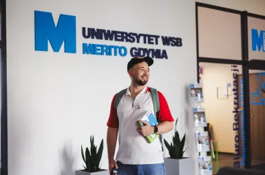 Zapraszamy na sesję, ale spokojnie – tylko zdjęciową! | Aktualności | Uniwersytet WSB Merito Gdynia: Student na korytarzu przy logo Uczelni
