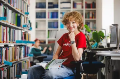 Zapraszamy na sesję, ale spokojnie – tylko zdjęciową! | Aktualności | Uniwersytet WSB Merito Gdynia: Studentka w bibliotece z książką na kolanach